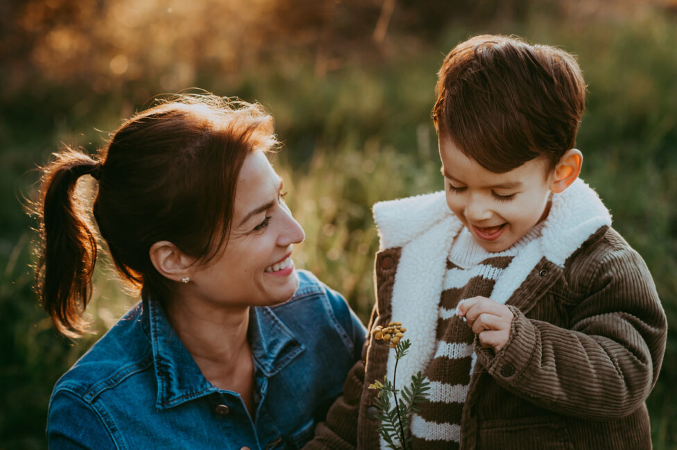 Mama zeigt ihrem Sohn auf einer herbstlichen Wiese im Nachmittagslicht eine Blume.