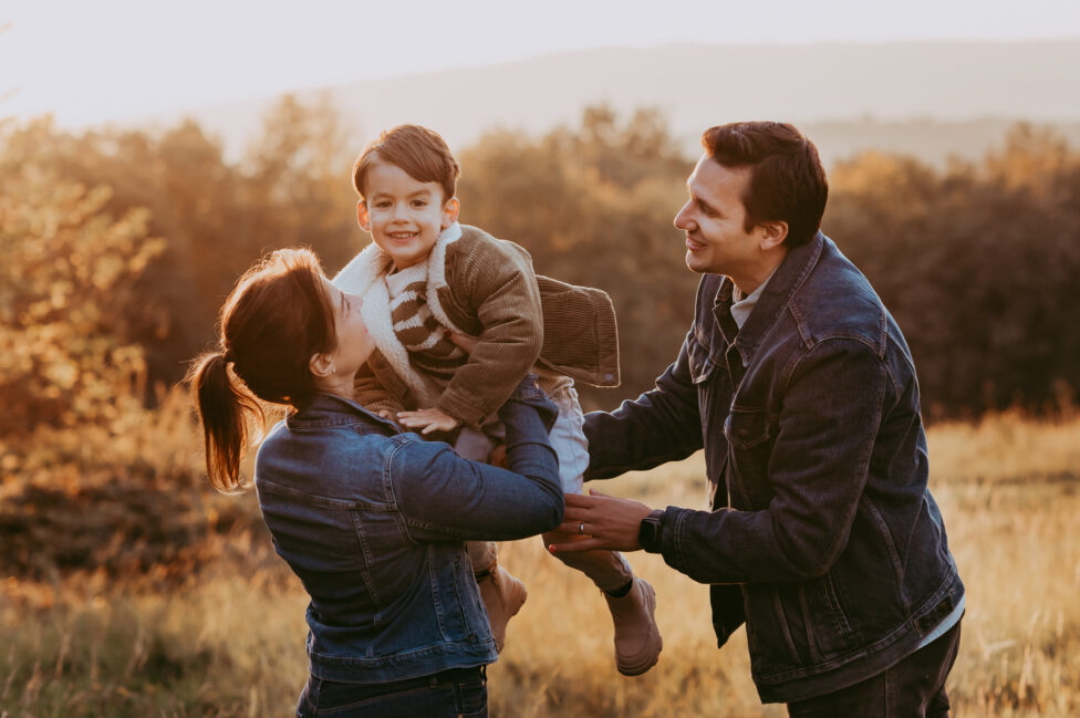 Familienportrait auf einer Wiese, Eltern tragen ihr Kind, sind fröhlich. Die Natur strahlt in herbstlichen Farben.